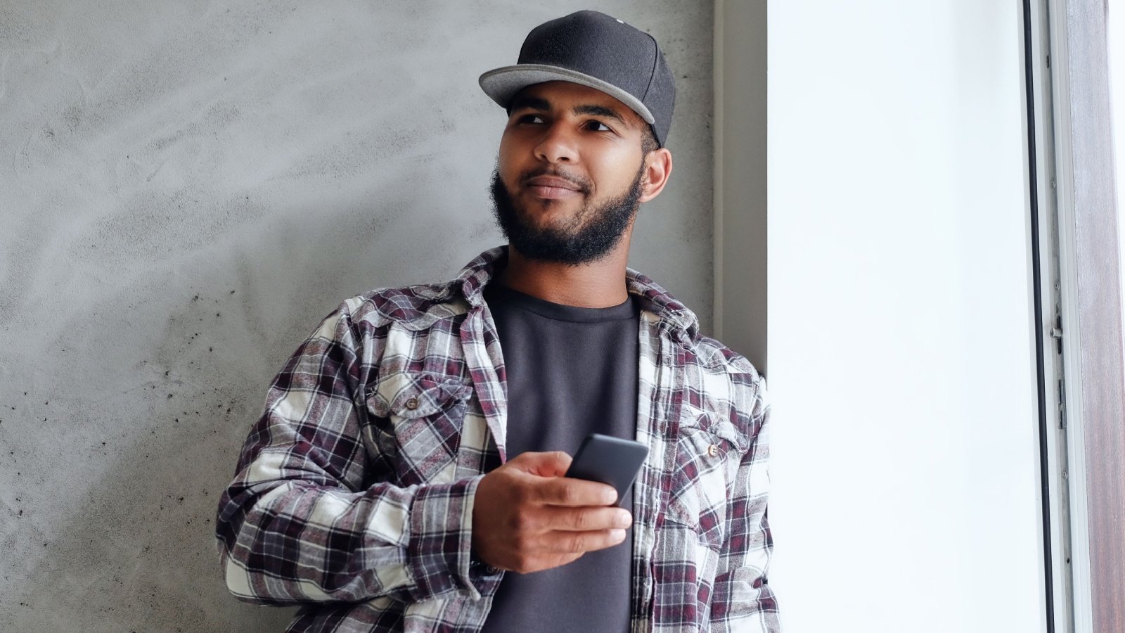 A black hipster male in a plaid fleece shirt and baseball cap hat sits near window smartphone FXQuadro Shutterstock