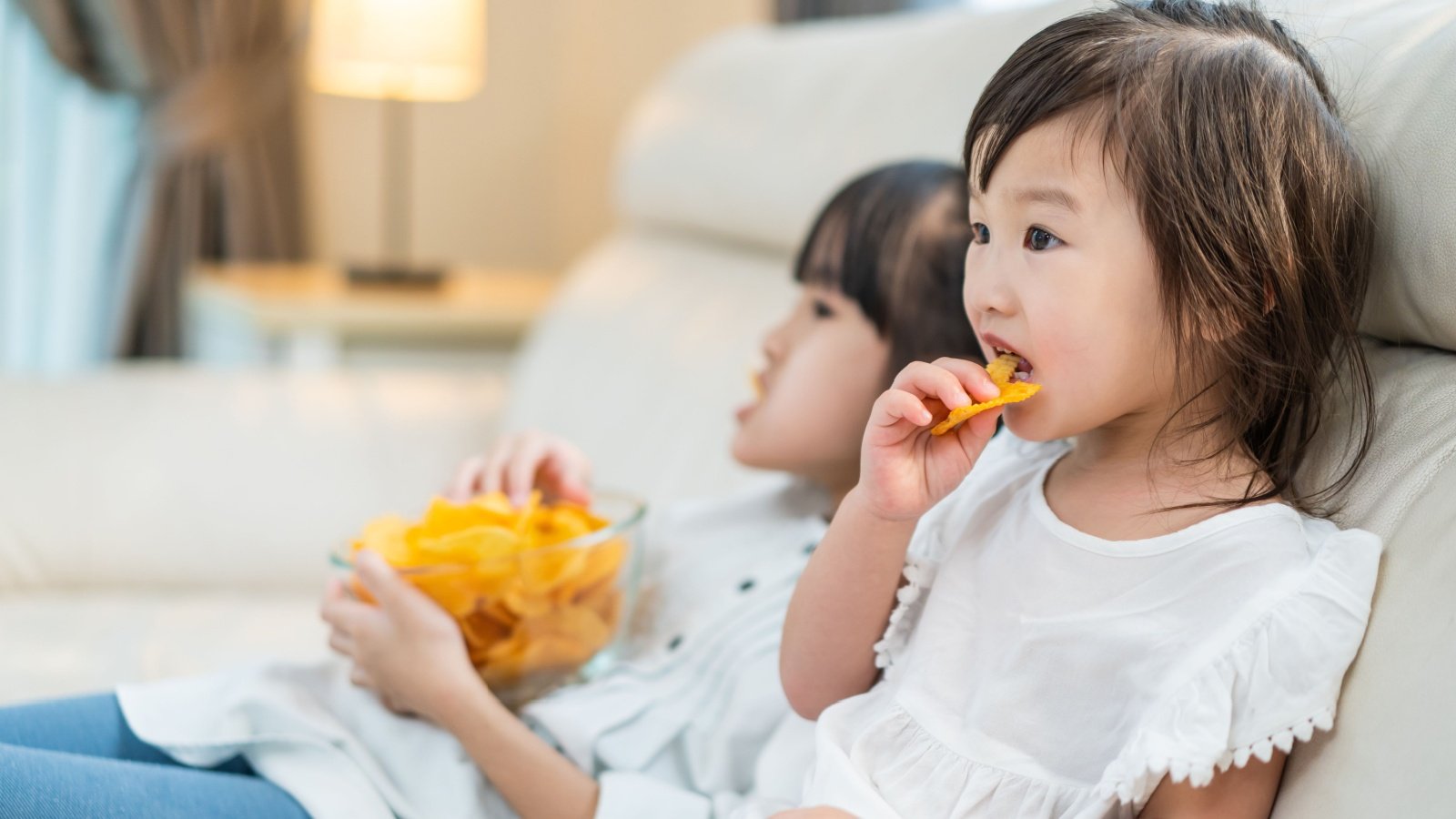 Asian Hungry child kid little girl sibling sisters Puts snack in mouth eat junk food couch potato chip watch tv movie Hananeko Studio Shutterstock