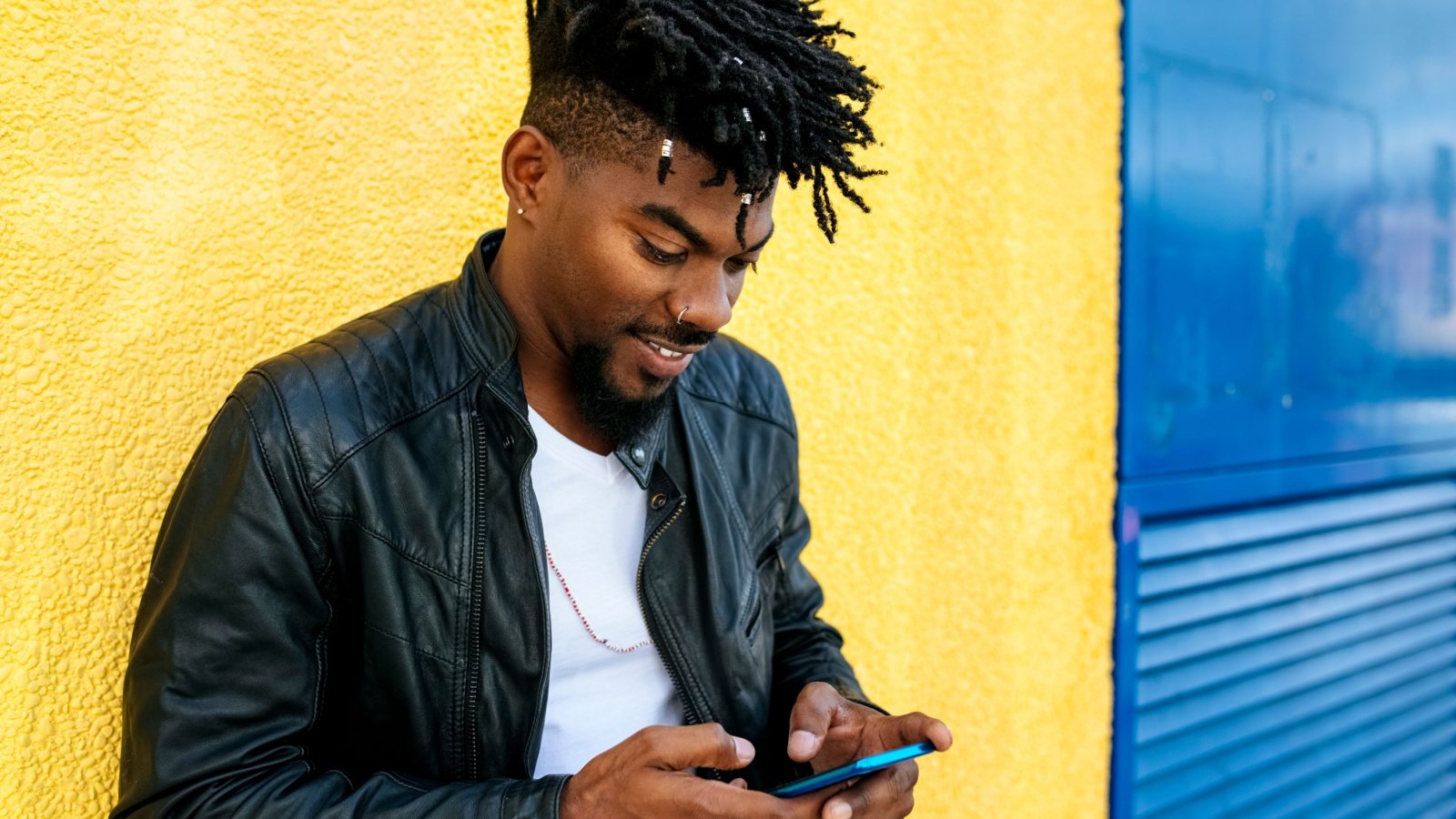 Black African American male leans against wall using mobile phone texting social media locs natural hair leather jacket TunedIn by Westend61 Shutterstock