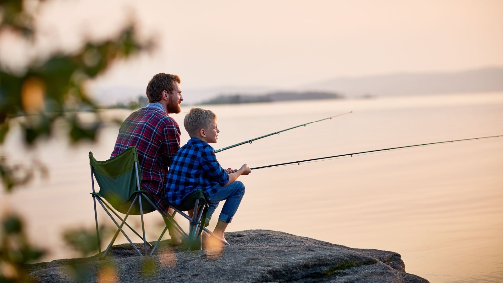 father and son sitting together on rocks fishing with rods in calm lake waters outdoor family fun Pressmaster Shutterstock