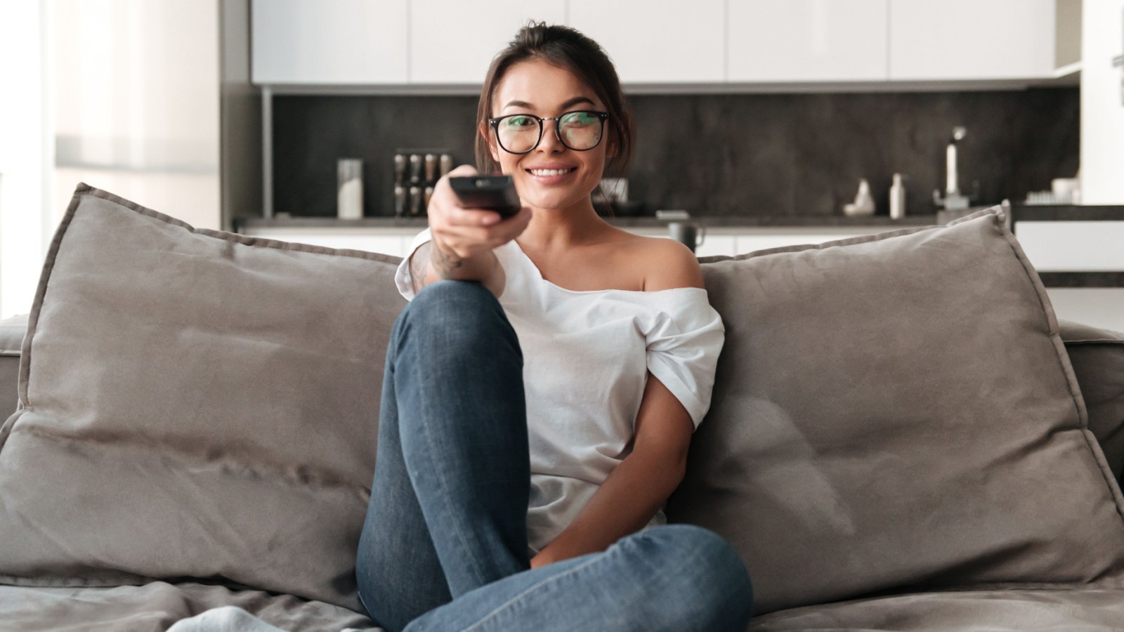 happy young woman sitting on sofa at home. Looking camera holding remote control watch TV Dean Drobot Shutterstock