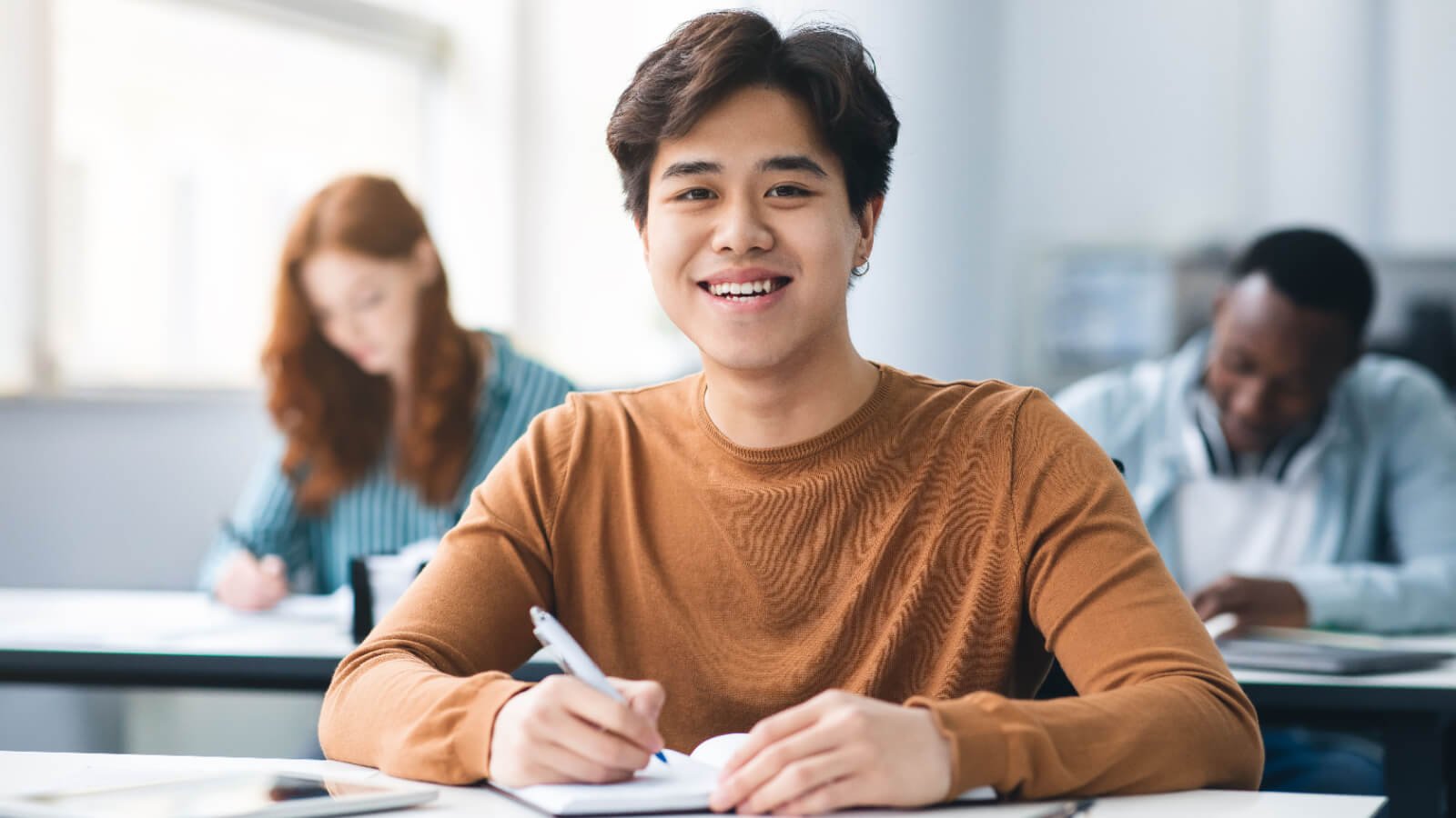 smiling asian male student sitting at desk in classroom at university taking test or writing notes prostock studio shutterstock